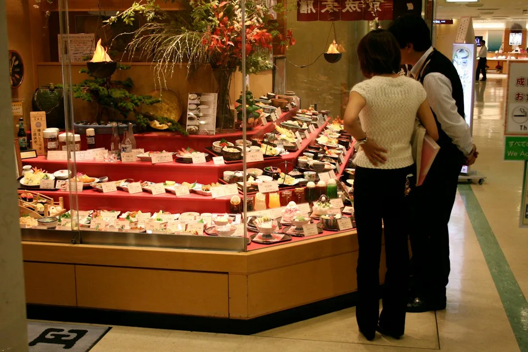 Bir Japon büyük mağazasının iç katında, fiyat etiketli yemek tabaklarıyla dolu cam vitrine bakan bir çift